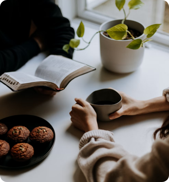 a photo of two people sitting together at a table. one is holding a drink and the other a book. They have a pot plant and some biscuits next to them